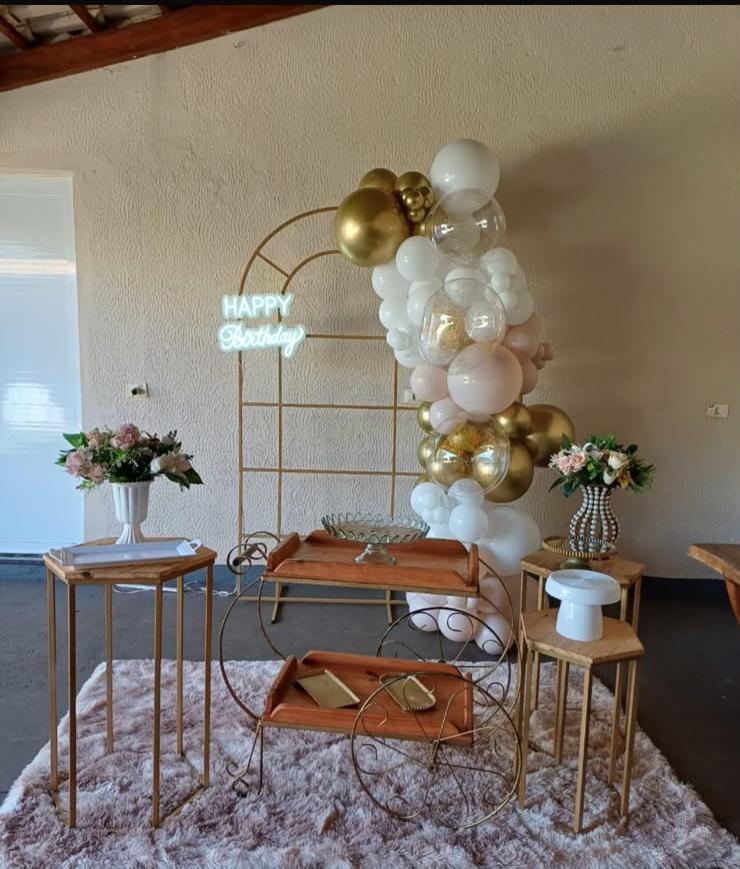 Cenário decorativo com arco dourado, mesas de apoio e arranjos florais assinado pela Boutique dos Balões